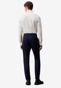 Uomo che indossa una camicia bianca a maniche lunghe, pantaloni eleganti blu scuro e scarpe eleganti nere, in piedi di spalle su uno sfondo neutro.