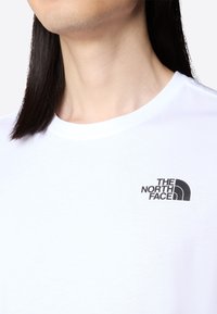 Close-up of a person wearing a white crew-neck t-shirt with a small black The North Face logo on the chest.