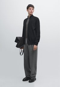 Zwarte cardigan met ritssluiting, daaronder een grijs T-shirt, grijze op maat gesneden broek, zwarte leren schoenen, en een zwarte leren tas in de hand. Minimalistisch ontwerp.