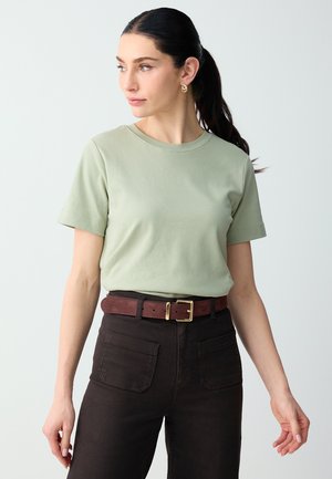Frau mit dunklem Pferdeschwanz trägt ein hellgrünes kurzärmeliges Shirt, dunkle Hosen mit Fronttaschen und einen braunen Gürtel mit goldener Schnalle, schaut nach links.