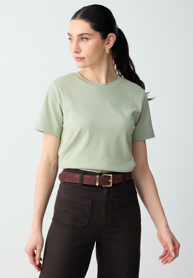 Femme avec queue de cheval foncée porte une chemise à manches courtes vert clair, pantalon foncé avec poches avant et ceinture marron avec boucle dorée, regardant vers la gauche.