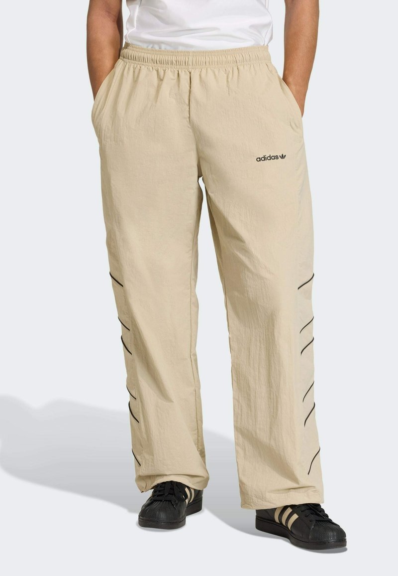 Pantaloni beige leggeri con vita elastica, tasche laterali e dettagli a strisce nere lungo le gambe. Logo Adidas sul davanti.