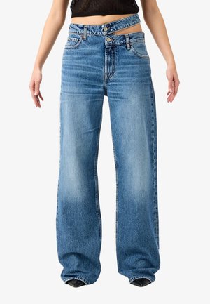 Donna che indossa jeans blu a vita alta e a gamba larga con due cinture sovrapposte e chiusura doppia con bottoni, abbinati a un top nero.