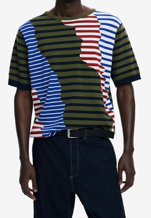 Homme portant un pull à manches courtes avec des motifs de rayures déchirées en vert olive, bleu marine, rouge, blanc et bleu, associé à un pantalon en denim foncé et une ceinture noire.
