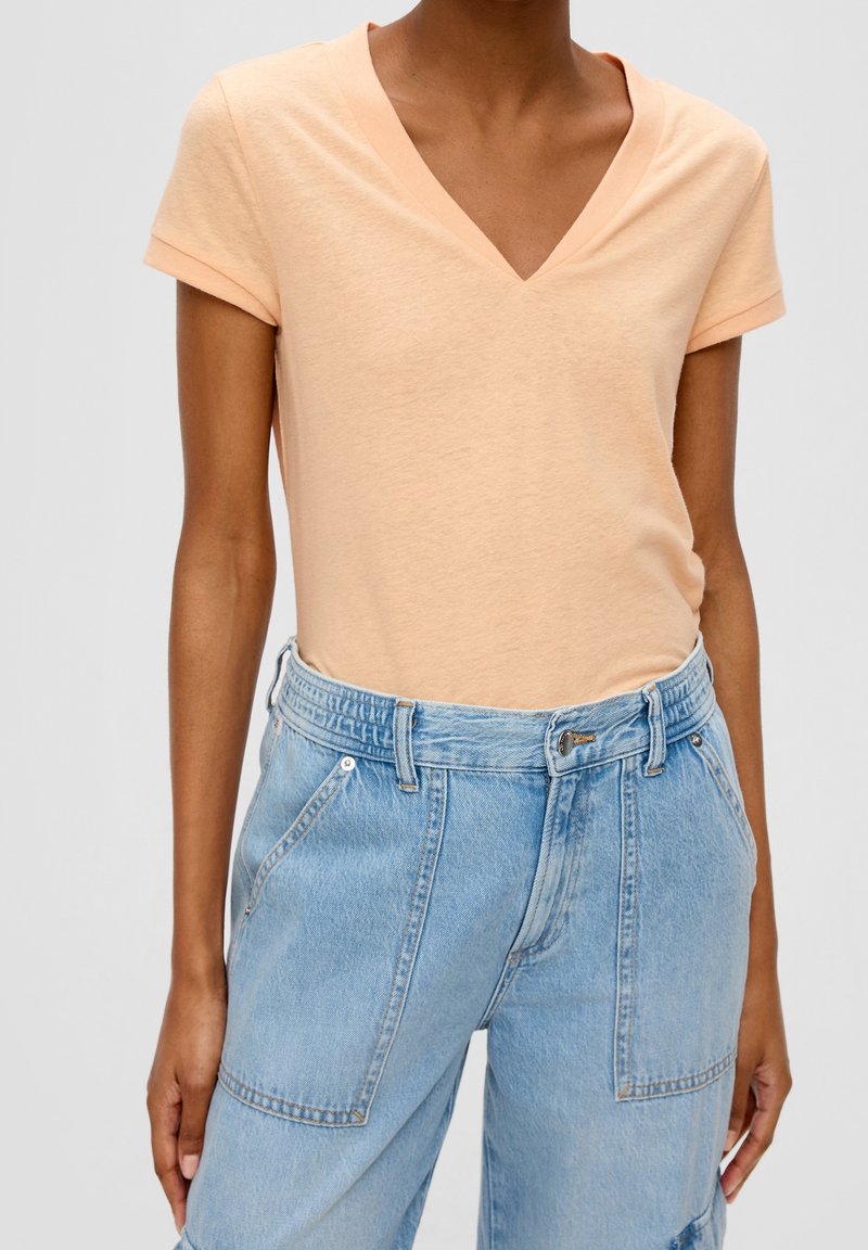 Personne portant un haut à manches courtes de couleur pêche claire avec un col en V, rentré dans un jean en denim bleu clair à taille haute avec des poches plaquées à l'avant.