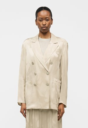 Vrouw die voorover staand naar voren kijkt, gekleed in een oversized beige double-breasted blazer en bijpassende rok tegen een effen achtergrond.