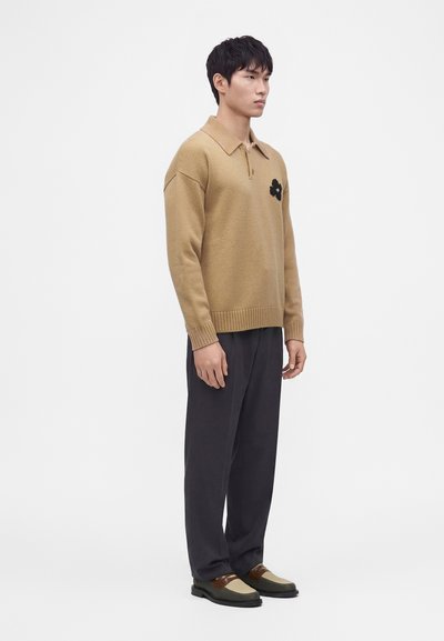 Homme portant un pull tricoté beige avec un détail de fleur noire, un pantalon sombre ample et des mocassins tissés bicolores, debout devant un fond blanc.