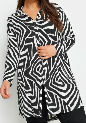 Woman wearing a long-sleeve black and white button-up shirt with abstract maze-like pattern, holding one hand near the buttons.