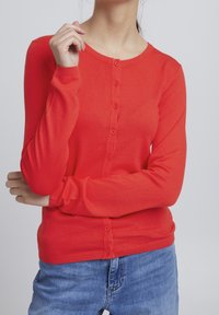 Cárdigan rojo de manga larga hecho de tela de punto, con un escote redondo, cierre de botón y puños acanalados, combinado con jeans azules.