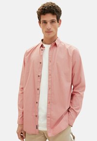 Rot-weiße gemusterte Button-Down-Hemd mit Kragen, langen Ärmeln und braunen Knöpfen. Über einem weißen T-Shirt getragen, mit sichtbaren Nähdetails.