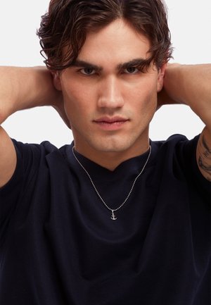 Silver anchor pendant on a beaded chain worn over a navy t-shirt. The model has short, wavy hair and a confident expression.