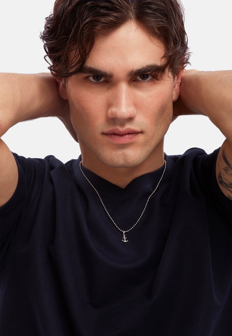 Sølv anker vedhæng på en perlekæde båret over en navy t-shirt. Modellen har kort, bølget hår og et selvsikkert udtryk.