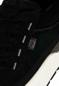 Sneaker in suede nero con finitura testurizzata, lacci neri, cuciture bianche e un piccolo tag marchiato sul lato con suola a contrasto.