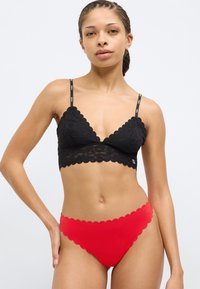 Bralette de encaje negro con bordes festoneados, tirantes finos con logo, combinado con bragas rojas con borde festoneado. Textura suave, diseño contemporáneo.