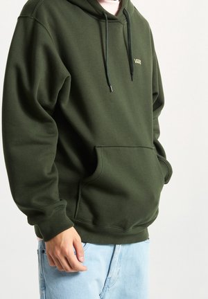 Personne portant un sweat à capuche Vans vert olive avec poche frontale et cordons noirs, assorti à un jean bleu clair, sur un fond uni.