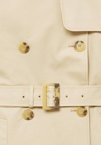 Gros plan sur un trench-coat beige avec de gros boutons dorés, une large ceinture avec une boucle rectangulaire dorée, et une texture de tissu lisse.