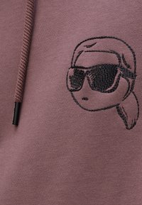 Bluza cu glugă de culoare mauve heathered, cu un personaj brodat texturat în negru purtând ochelari de soare, având șnur și margini cu nervuri.