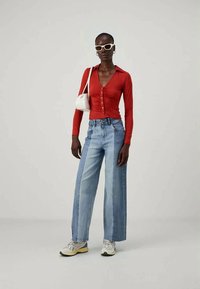 Top ajustado de mangas longas vermelhas com detalhes em botão, combinado com jeans de corte largo em dois tons. Acessórios incluem uma bolsa branca e óculos de sol.