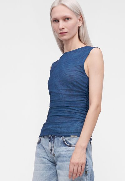 Femme aux cheveux blonds platine raides, portant un haut bleu froncé sans manches et un jean bleu clair, debout devant un fond blanc uni.