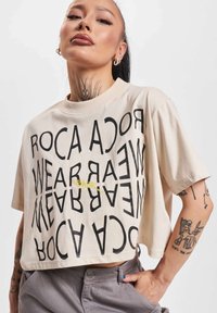 Beige cropped t-shirt with bold black text arranged vertically and horizontally, featuring a small yellow accent. Soft cotton fabric.