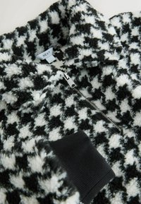 Fleece cu model houndstooth alb și negru, cu fermoar, având o suprafață moale și texturată. Prezintă un manșon negru cu nervuri și un fermoar argintiu. Eticheta brandului este vizibilă.