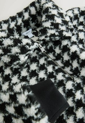 Mustavalkoinen houndstooth-kuvioinen vetoketjullinen fleece, jossa on pehmeä, teksturoitu pinta. Varustettu mustalla ribbikujalla ja hopeisella vetoketjulla. Brändilogo näkyvissä.