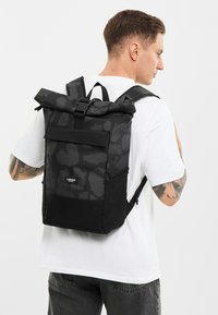 Rolový batoh v čiernej farbe s matným povrchom, s jemným kamuflážnym vzorom, nastaviteľnými popruhmi a prednou kapsou s logom značky.