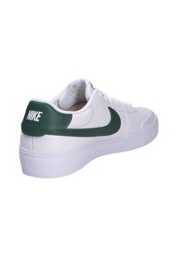 Zapatilla Nike blanca con acentos verdes; presenta un upper sintético liso, talón texturizado y suela de goma. Diseño clásico de tipo bajo.