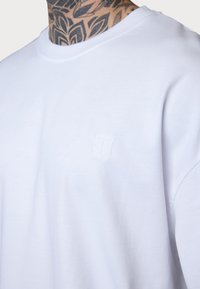 White cotton sweatshirt with a crew neckline, featuring a small embroidered logo on the chest. Soft texture, relaxed fit, minimal design.