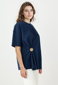 Marineblaues T-Shirt aus weichem Stoff, mit einem gedrehten Design und einem goldenen Knopf an der Taille, kurzen Ärmeln und rundem Halsausschnitt.