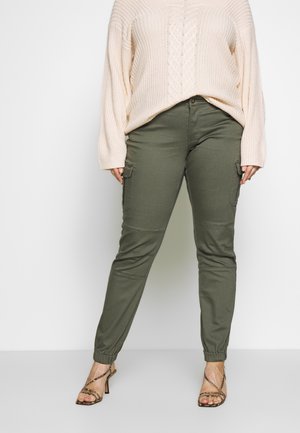 Frau trägt einen beigefarbenen Zopfstrickpullover, olivgrüne Cargohosen mit Beinsaum und schlangenprintbesetzte Riemchen-Sandaletten mit hohen Absätzen.