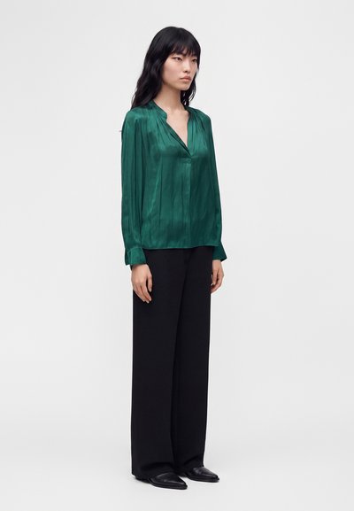 Femme aux longs cheveux noirs portant un chemisier en satin vert profond, un pantalon noir à jambes larges et des chaussures noires à bout pointu, debout devant un fond uni.