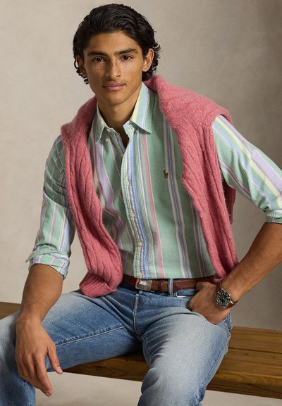 Mladić tamne kose, odjeven u zelenu prugastu košulju, ružičasti džemper preko ramena, plave traperice i ručni sat, sjedi na klupi.