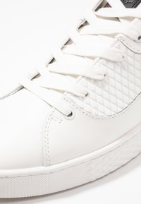 White sneaker with a smooth leather upper, textured side panel, and flat laces. Features a textured rubber sole with a diamond pattern.