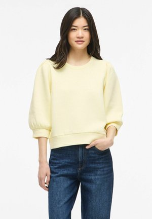 Femme aux longs cheveux foncés portant un sweat-shirt jaune clair à manches bouffantes et un jean bleu, debout avec une main dans la poche sur fond blanc.