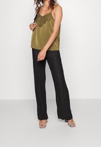 Olijfgroene satijnen camisole met kantafwerking, gecombineerd met zwarte geplooide wijde broek en open tenen, zwart-wit gestreepte hakken.