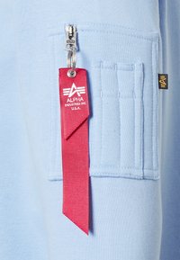 Světle modrá mikina s zipem a výraznou červenou štítkem s bílým logem. Obsahuje texturovanou náprsní kapsu s vertikálními pruhy.