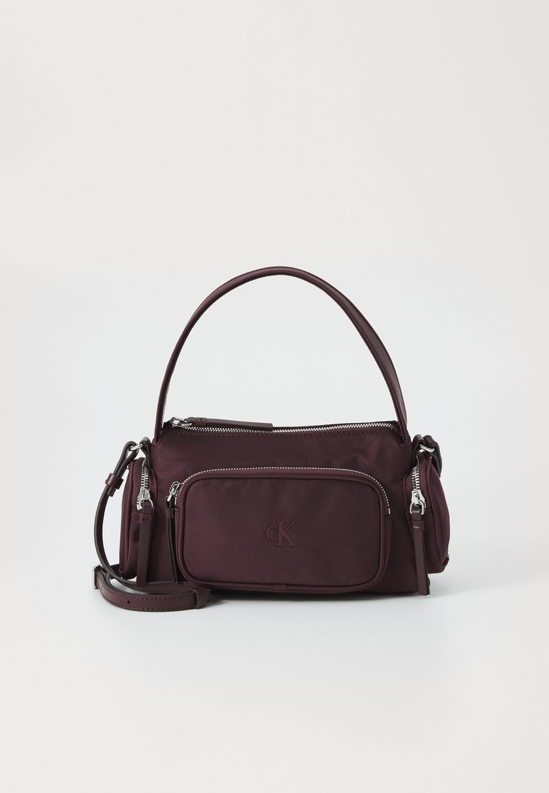 Sac à main Calvin Klein violet foncé avec poignée supérieure, poche avant zippée, détails zippés sur les côtés et bandoulière réglable sur fond blanc.