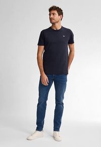 Marinblå t-shirt med rund hals, kombinerad med ljusblå jeans och vita sneakers, stående mot en minimalistisk bakgrund.