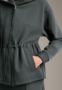 Tmavozelená zipová bunda vyrobená z hladkej tkaniny, s gumičkovým pásom v páse, bočnými vreckami a ribbovanými manžetami. Jednoduchý, moderný dizajn.