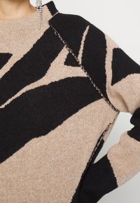 Pull tricot beige et noir avec motif géométrique abstrait et coutures visibles à l’épaule et à la manche.