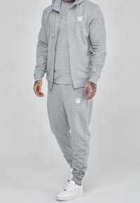 Chándal gris hecho de una tela suave y texturizada, que presenta una sudadera con cremallera, mangas largas y puños elásticos; combinado con zapatillas blancas.