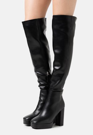 Botas de cuero negro hasta la rodilla con tacones altos y gruesos, y cremalleras laterales, usadas sobre piernas desnudas.