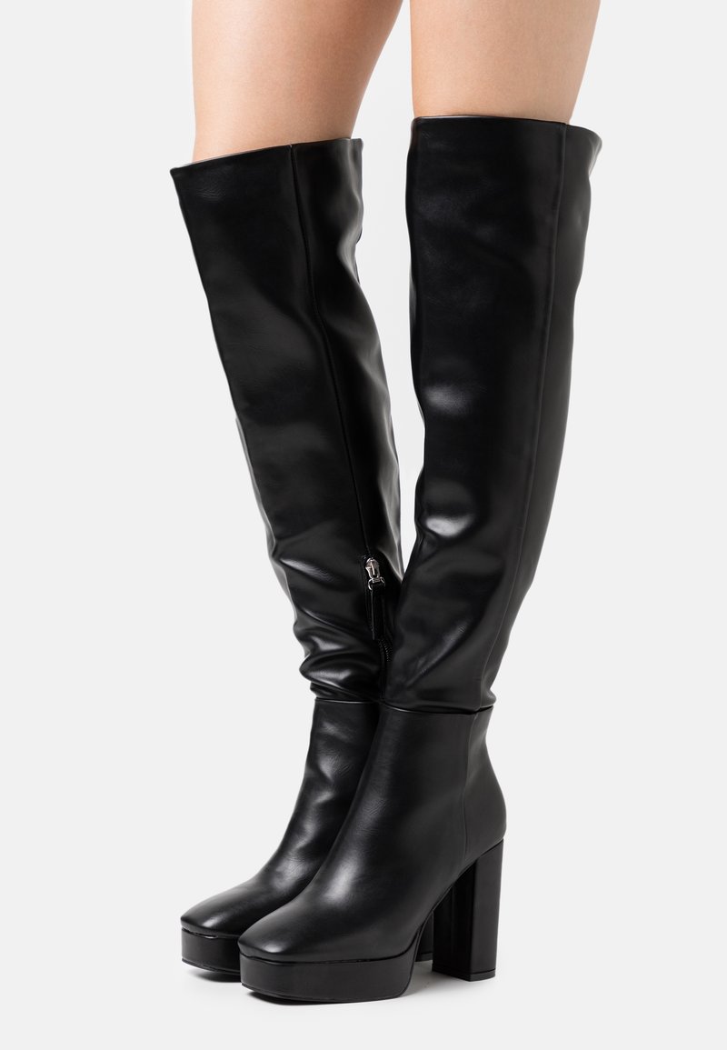 Botas de cuero negro hasta la rodilla con tacones altos y gruesos, y cremalleras laterales, usadas sobre piernas desnudas.