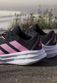 Para czarno-różowych butów do biegania Adidas z białymi podeszwami Cloudfoam umieszczona na zewnątrz na chodniku obok roślin.