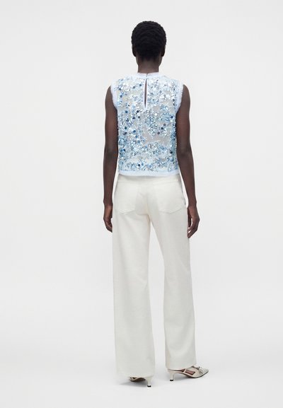 Άτομο που φοράει ένα αμάνικο διάφανο τοπ με μπλε κεντήματα λουλουδιών και λευκό παντελόνι με φαρδιά πόδια, στέκεται πλάτη σε μονόχρωμο φόντο.