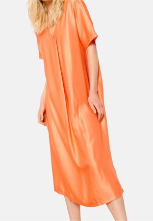 Frau trägt ein locker sitzendes, glänzendes oranges Midikleid mit kurzen Ärmeln und leichtem V-Ausschnitt, steht vor einem schlichten hellen Hintergrund.