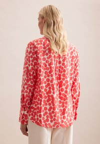 Blusa rossa e bianca con un motivo circolare, dotata di maniche lunghe e una vestibilità comoda. Il materiale sembra liscio con una leggera lucentezza.