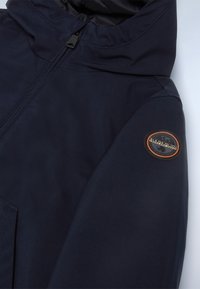 Navy modrá nepromokavá bunda s vysokým límcem, předním zipem a kulatou nášivkou na levém rukávu. Hladká textura, minimalistický design.