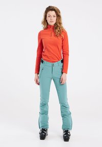 Felrood langwerpig shirt met hoge kraag, gecombineerd met teal sneeuwbroeken met ritszakken en zwarte accenten. Zwarte skibotten.